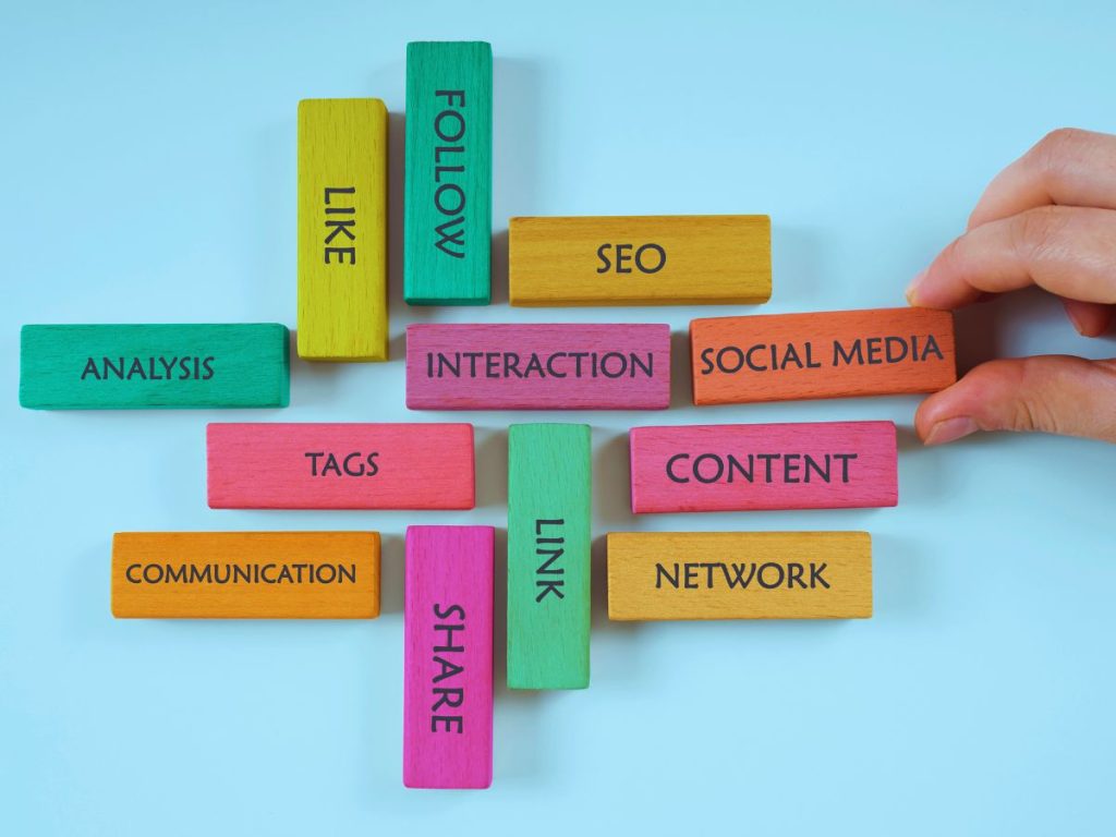 Colorful wooden blocks with social media-related words such as 'FOLLOW', 'LIKE', 'SEO', 'SOCIAL MEDIA', 'CONTENT', 'NETWORK', 'SHARE', 'COMMUNICATION', 'TAGS', 'INTERACTION', 'LINK', and 'ANALYSIS' arranged on a light blue background, with a hand picking up one of the blocks.
